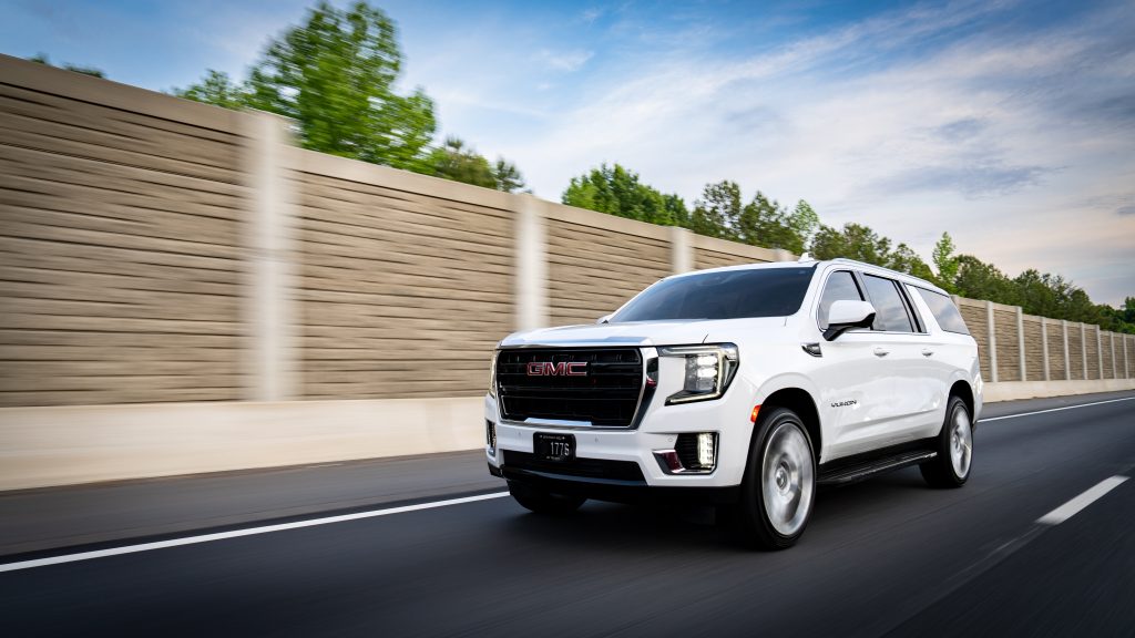White GMC Yukon SUV driving on a highway with a blurred brown concrete barrier and blue sky in the background, motion blur suggesting speed.