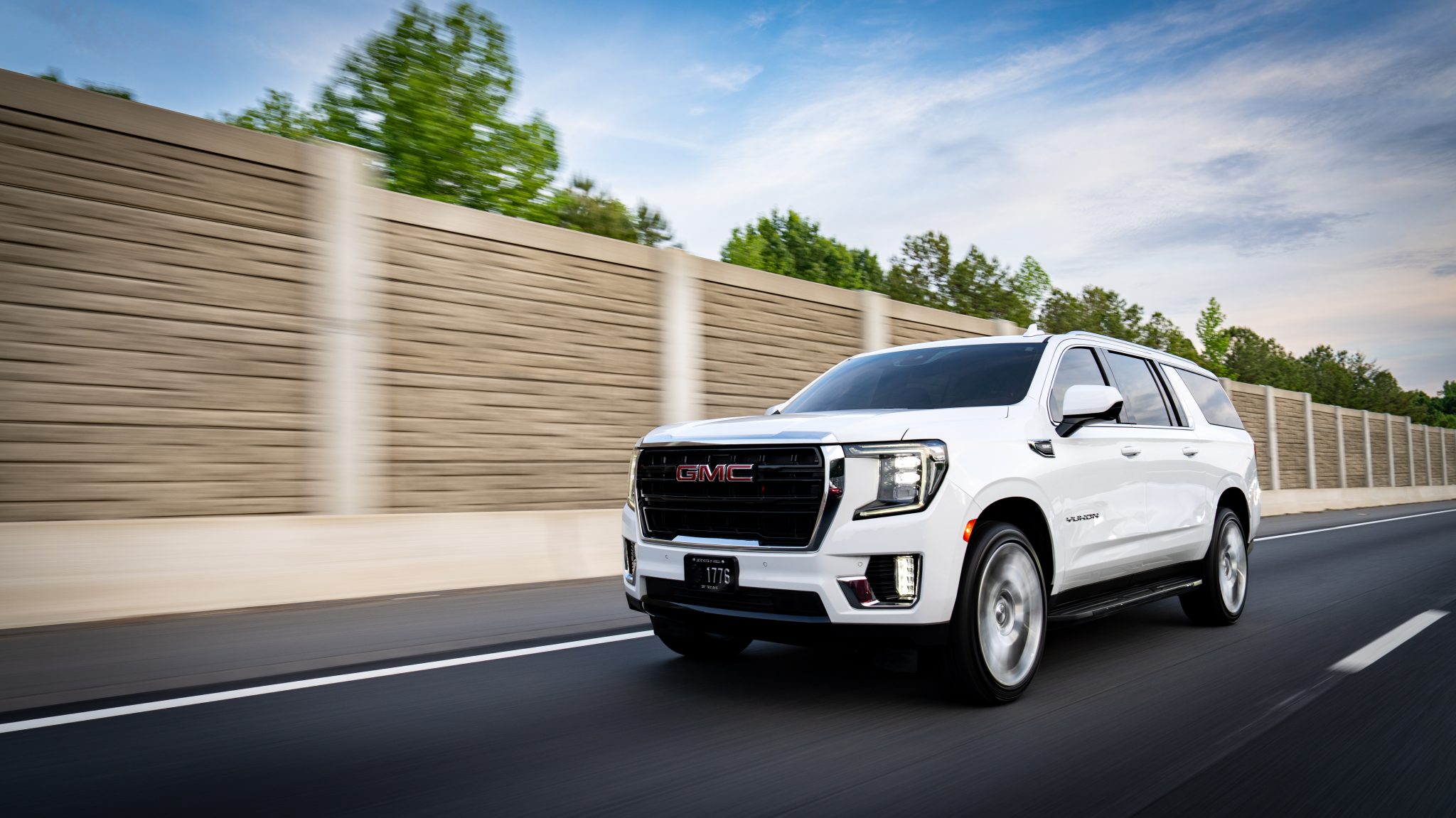 White GMC Yukon SUV driving on a highway with a blurred brown concrete barrier and blue sky in the background, motion blur suggesting speed.