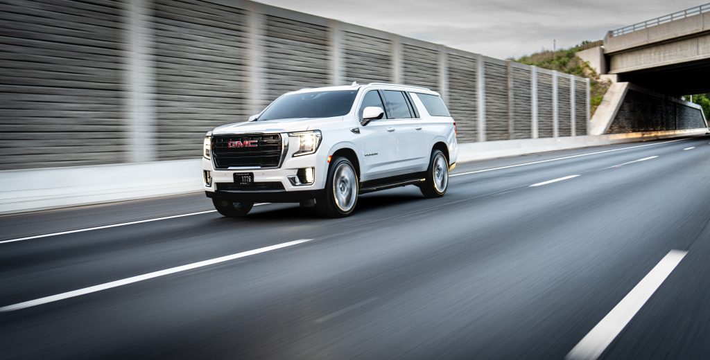 White GMC SUV in motion on a multi-lane highway beside barriers and a tunnel/overpass in the distance.