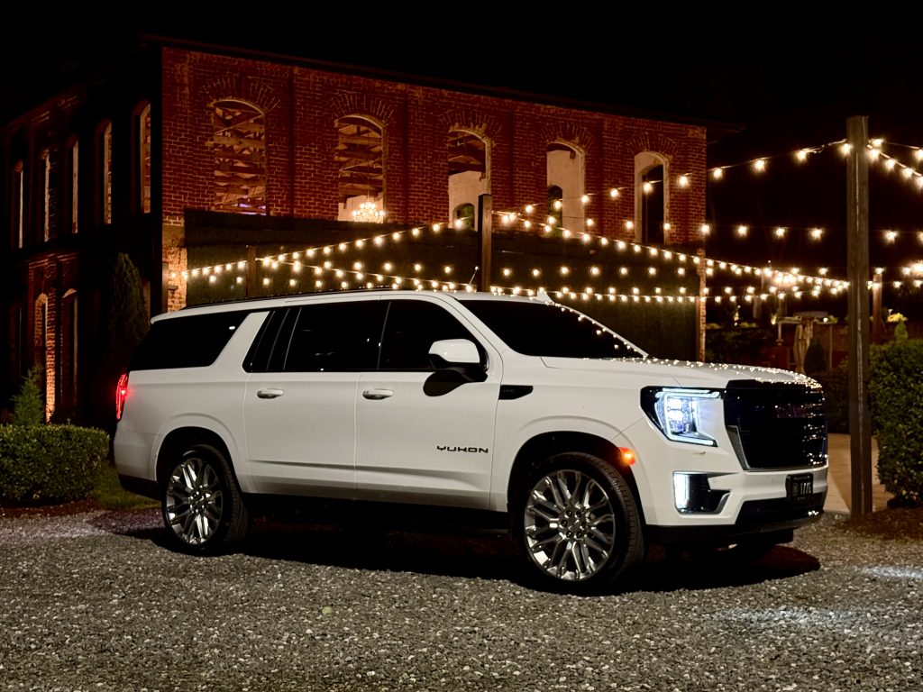 White SUV (Yukon) parked on gravel at night in front of a brick building with string lights overhead.