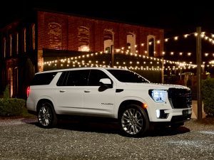 White SUV (Yukon) parked on gravel at night in front of a brick building with string lights overhead.
