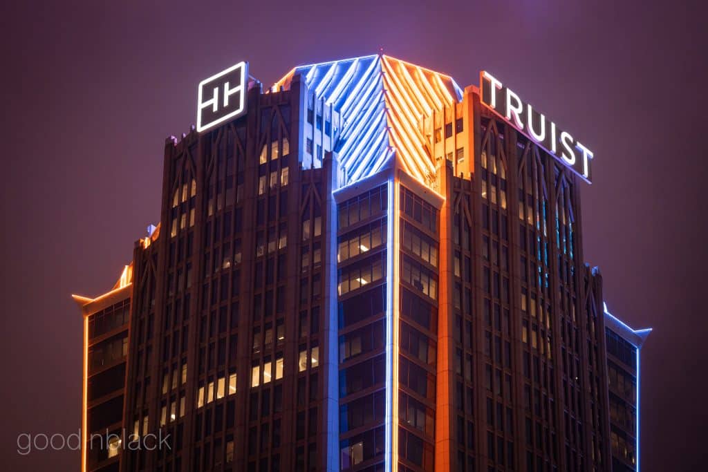 Nighttime view of a tall office building with blue and orange neon lighting along the roof and a white square logo on the corner. Charlotte, NC Truist Building